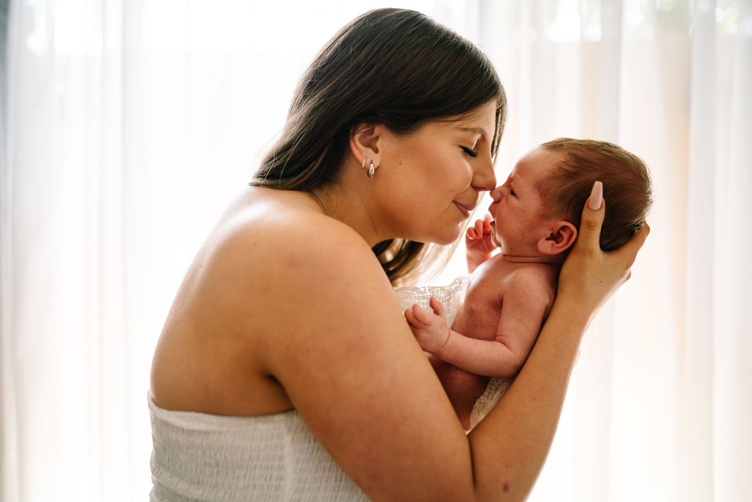 Mother in white tenderly holding and interacting with newborn baby against soft window light taken by Melbourne newborn photographer.