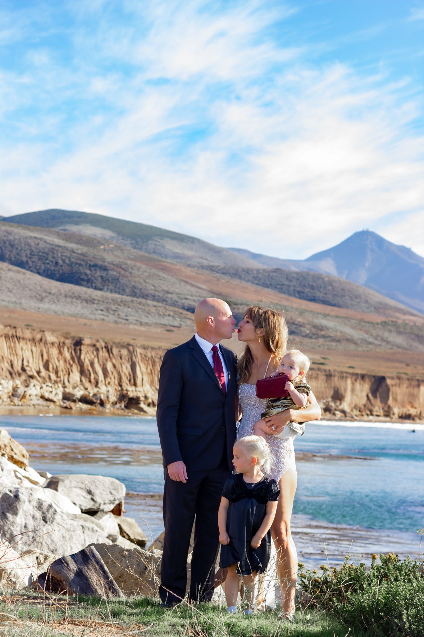 Family group poses for photos on rocky coastline with mountains and lake in background.