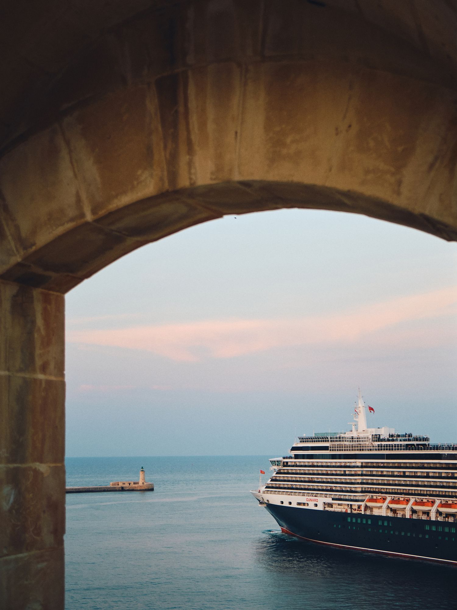 Cruise ship viewed through stone archway at sunset with lighthouse visible in calm blue ocean waters.