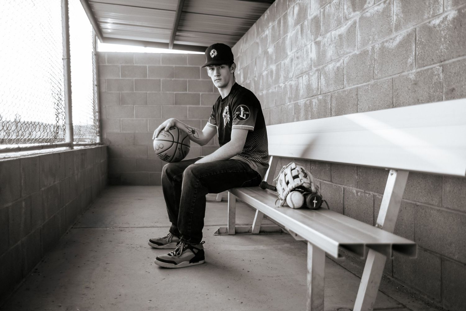 A baseball player sits alone on a bench in a concrete dugout holding sports equipment.