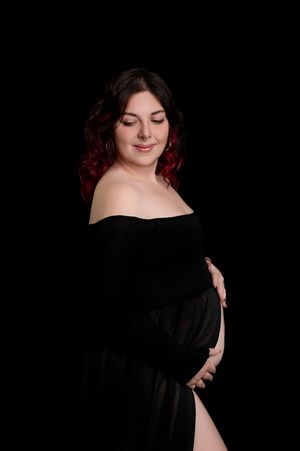 A maternity portrait of a person in a black off-shoulder dress against a dark background, eyes closed with a peaceful expression.
