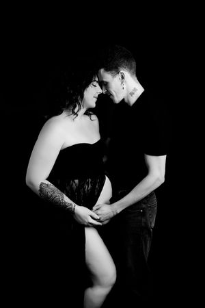 An intimate black and white maternity photo of a couple sharing a tender moment against a dark backdrop.