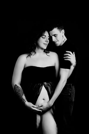Black and white intimate maternity photo of an expecting couple embracing against dark background.