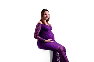 A pregnant woman in a full-length purple lace maternity gown sits elegantly against a white background.