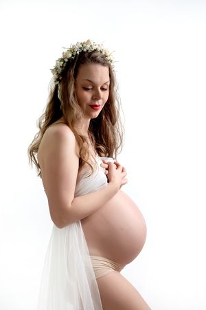 Ethereal maternity portrait with figure in white fabric and floral crown gazing down at baby bump against light background.