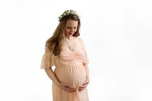 Expectant mother in peach-colored lace dress and floral crown gently cradling her baby bump against white background.