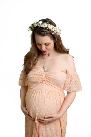 Expectant woman wearing peach lace dress and floral crown looks down at her belly in gentle maternity portrait.
