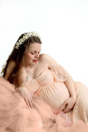 Expecting mother in a peach lace gown and floral crown poses gracefully against soft pink tulle in maternity photo.