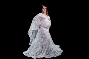 Person in long white lace maternity gown poses against dark background for elegant portrait.
