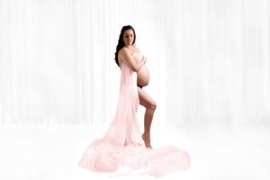Pregnant woman in flowing pink dress poses elegantly on white background for maternity photo shoot.