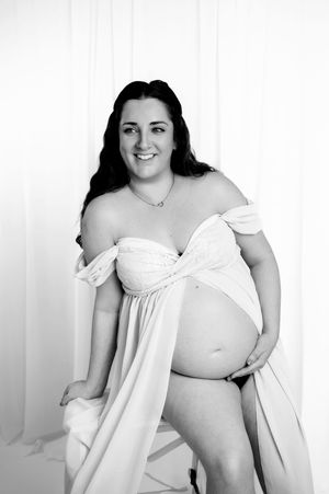 Monochromatic portrait shows an expectant mother in flowing white fabric against a light backdrop.