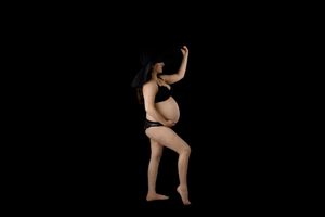 Pregnant woman in black underwear strikes a graceful dance pose against dark background in artistic maternity photo.