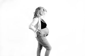 A pregnant figure in casual denim overalls and white shirt poses in black and white against a light backdrop.