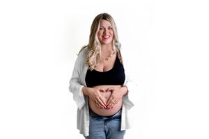 A pregnant person wearing a black tank top and grey cardigan makes a heart shape with hands on their belly.