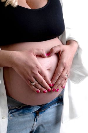Close-up of a pregnant belly with hands forming a heart shape, wearing black top and denim jeans.