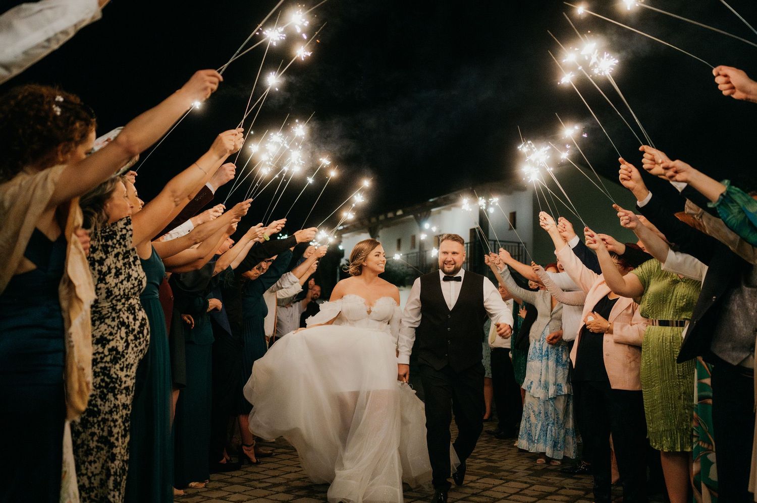 Wedding guests hold sparklers forming a tunnel as happy couple exits through glowing archway at nighttime celebration.