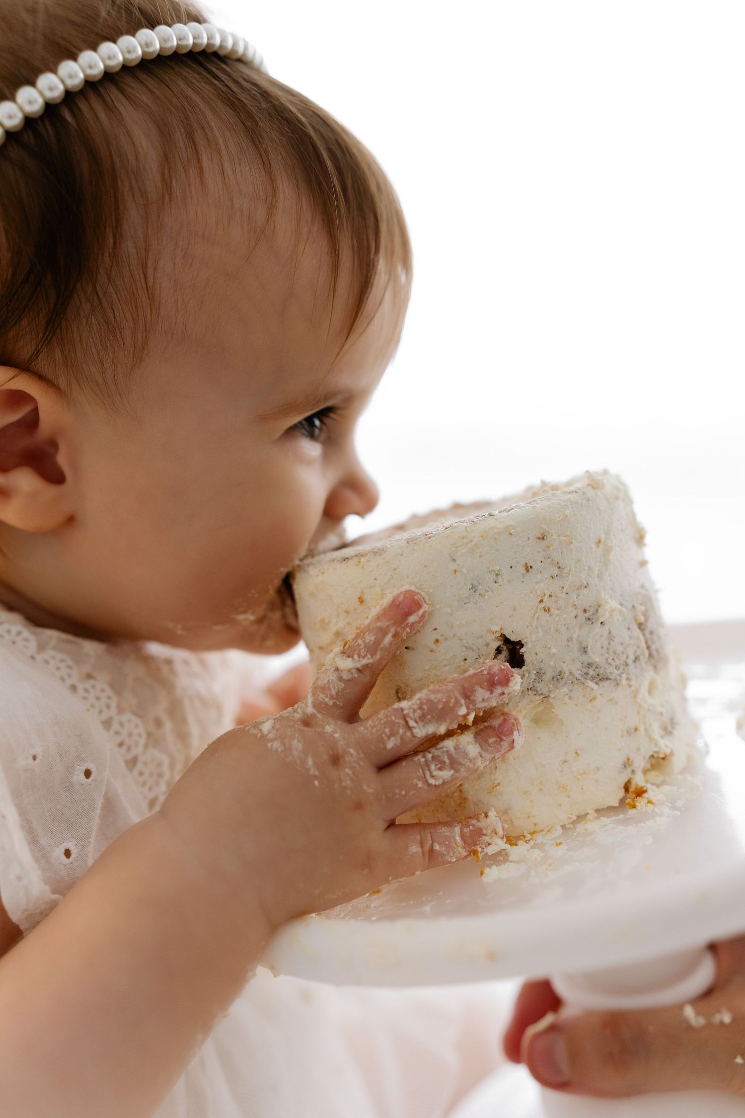 Small baby in white dress takes a big bite of cake while sitting at their first birthday celebration.
