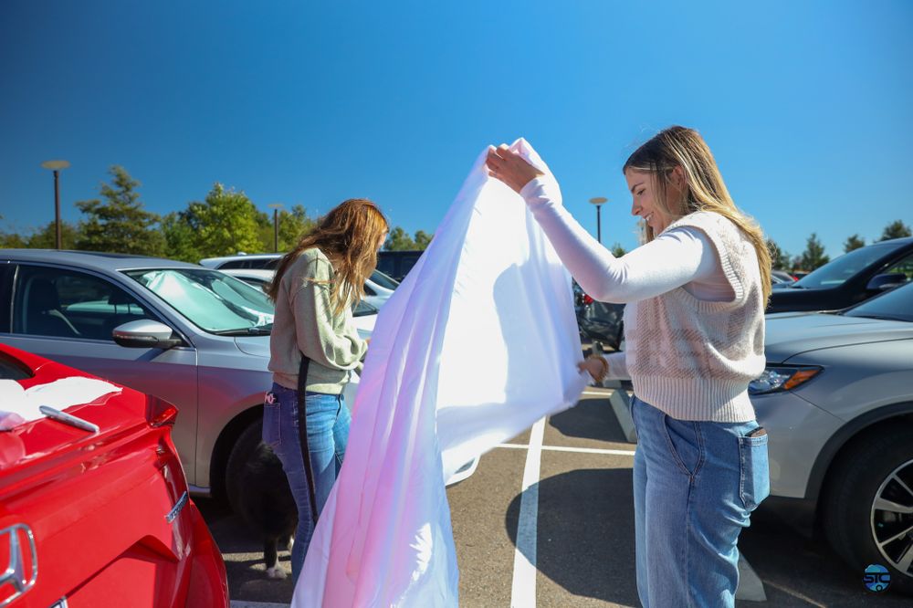 Two people examine a long white tulle fabric in a sunny parking lot surrounded by cars.
