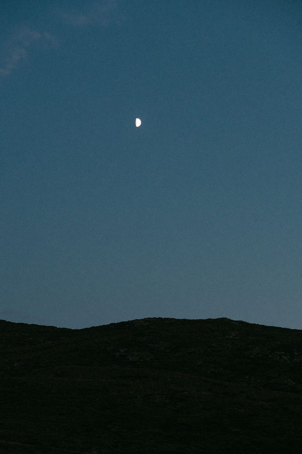 Half moon glowing in a dark blue sky above silhouetted mountain ridges. Destination wedding photographer Kythnos
