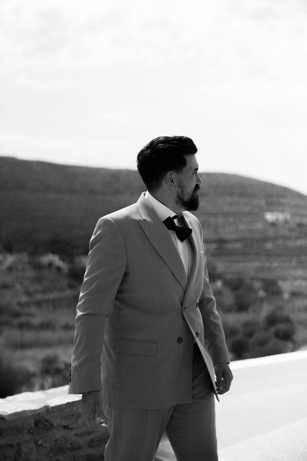 Groom in light gray suit stands against scenic hillside backdrop in black and white photograph. Destination wedding photographer Kythnos
