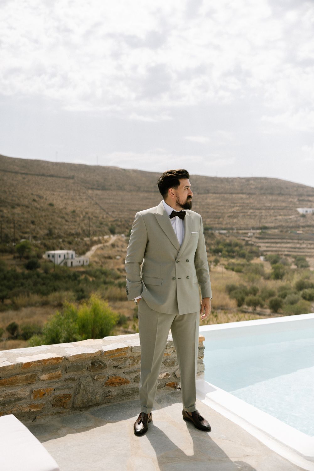 Groom in light suit and bowtie stands by infinity pool with mountain landscape. Destination wedding photographer Kythnos