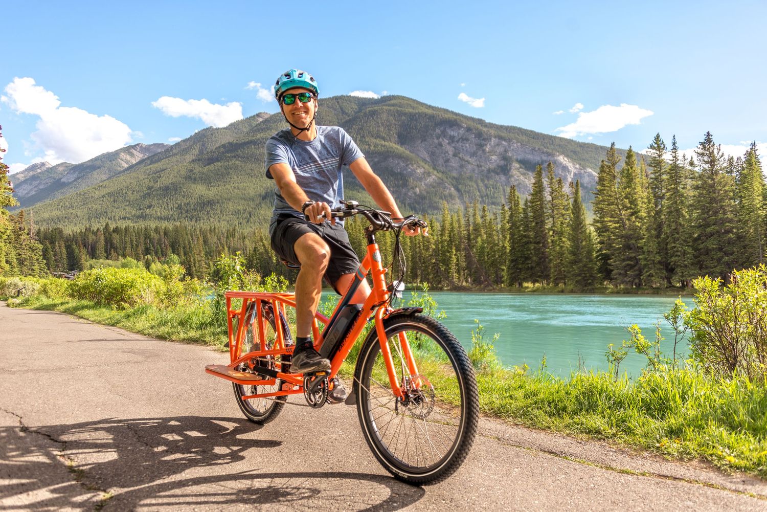 Cyclist riding orange bike on scenic path along turquoise river with mountain and forest landscape in background.