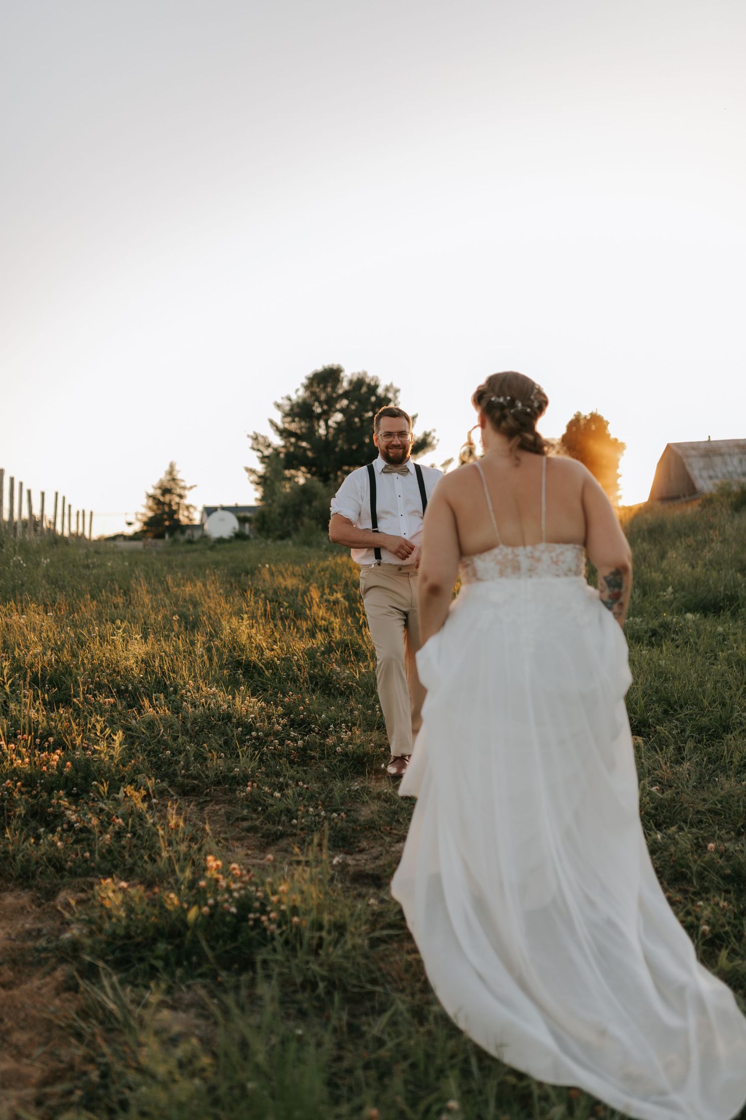 Groom smiling as bride walks toward him through wildflower field at sunset.