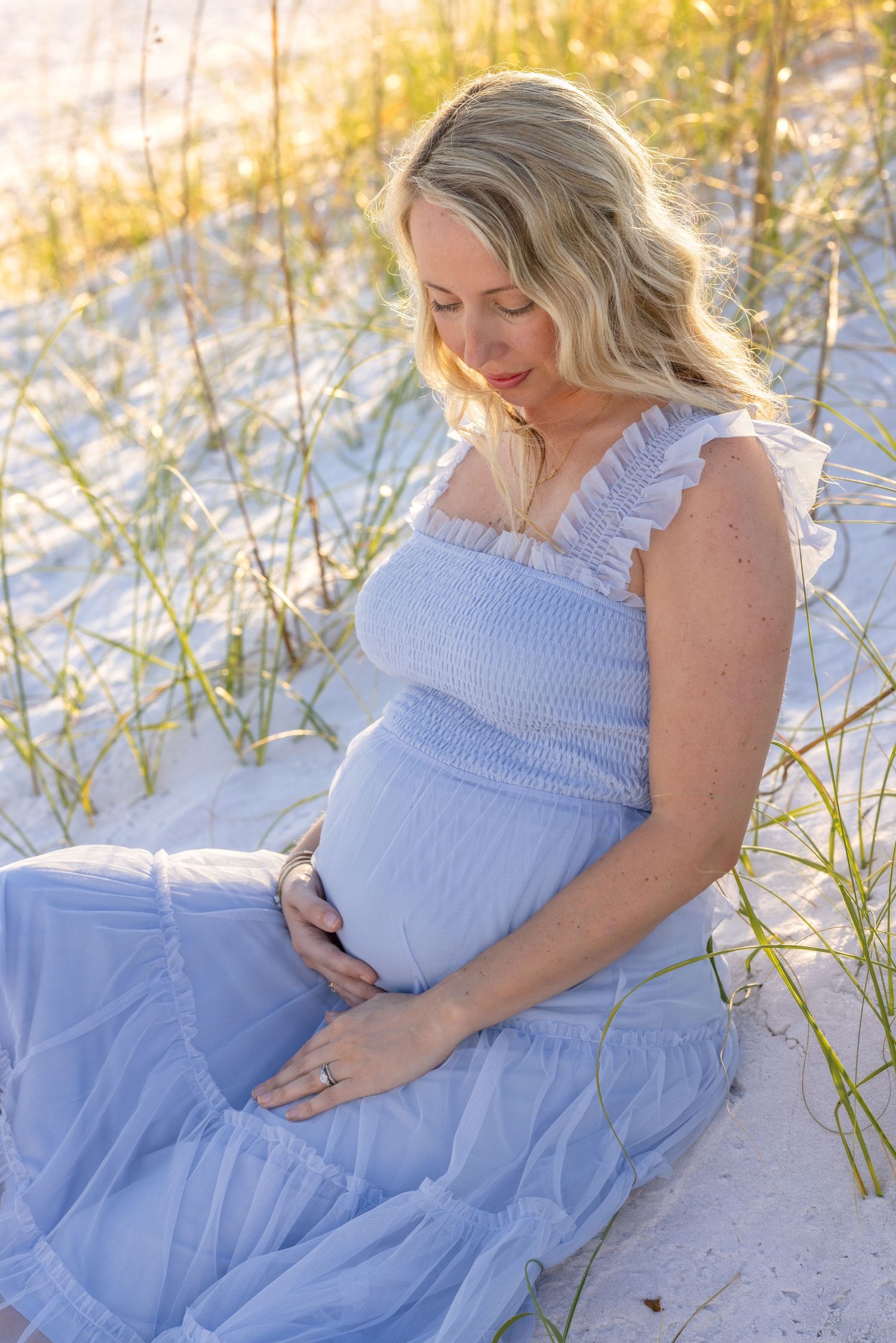 Expectant woman in flowing blue dress sits peacefully on sandy beach dunes during golden sunset.