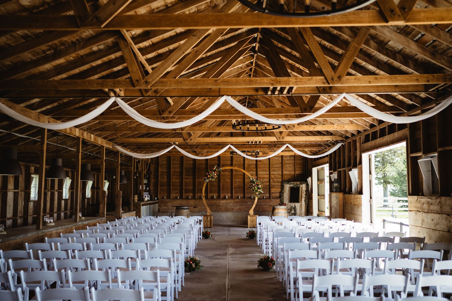 A rustic barn wedding venue interior with wooden beams, white fabric draping, and rows of white chairs set up for ceremony.