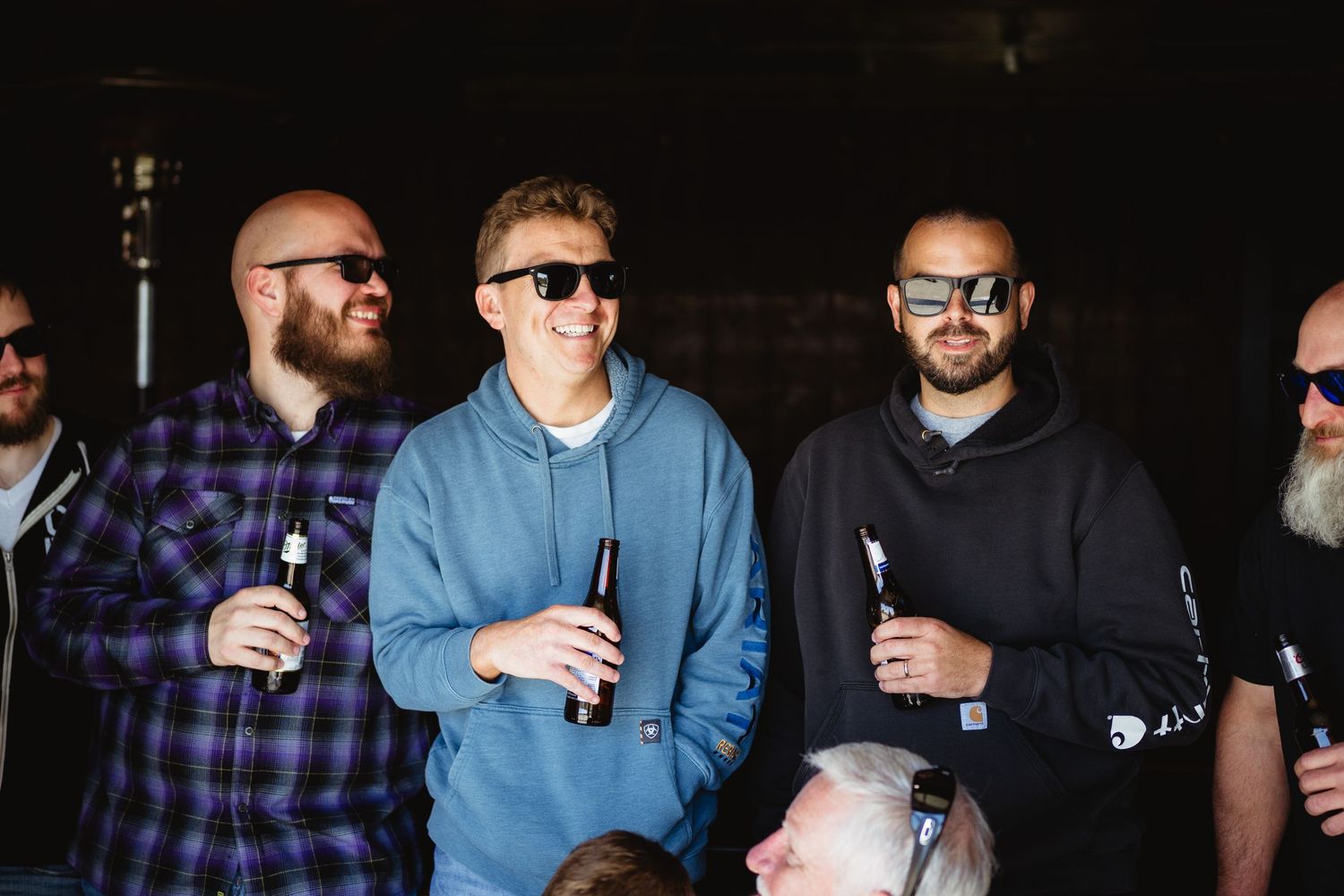 A group of friends enjoying drinks together while wearing casual attire and sunglasses on a dark background.