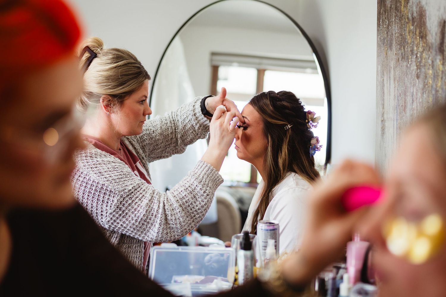 A makeup artist applies cosmetics to a woman while she sits in front of a large round mirror.