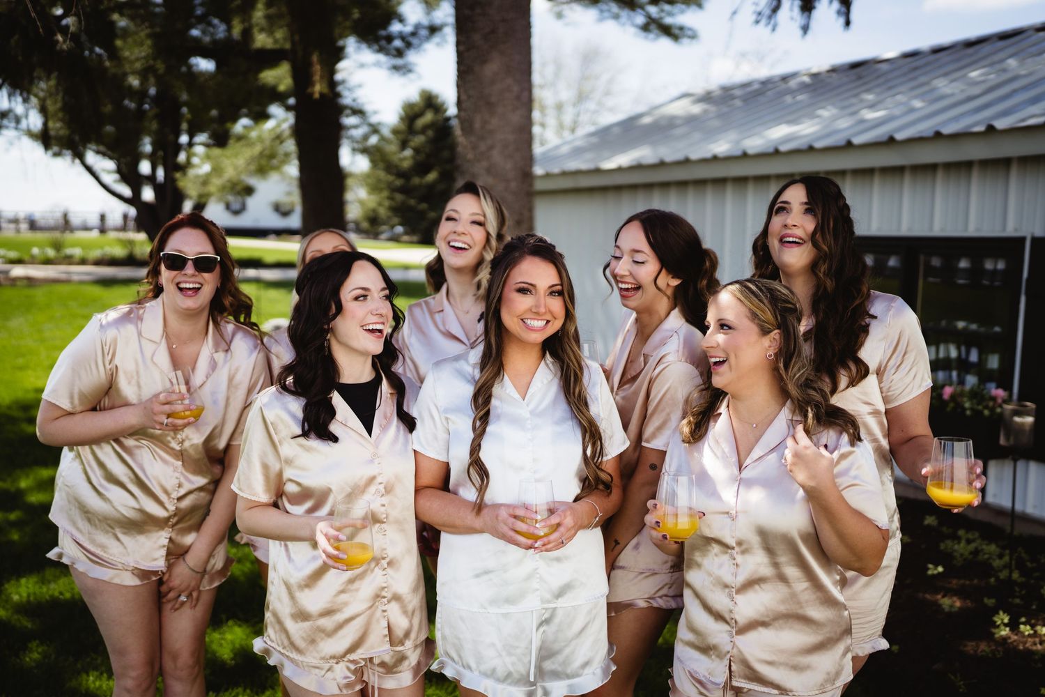 A group of friends in matching satin robes celebrate together outdoors on a sunny day near a rustic building.