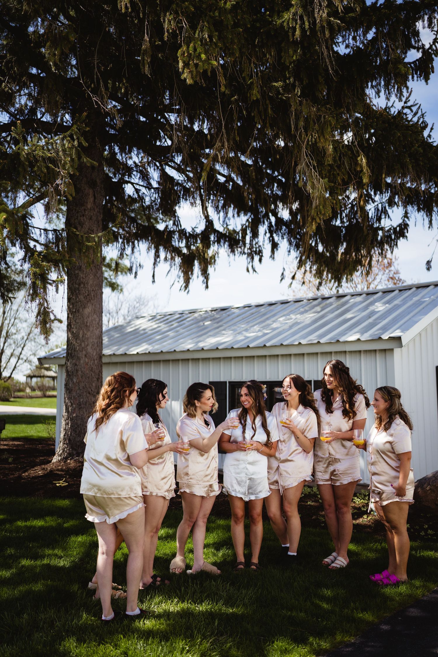 A group wearing matching white outfits stands in casual conversation near a white barn under an evergreen tree.