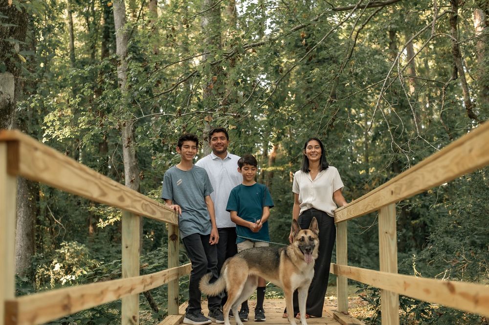 Family posing with their German Shepherd dog on wooden bridge in wooded setting.