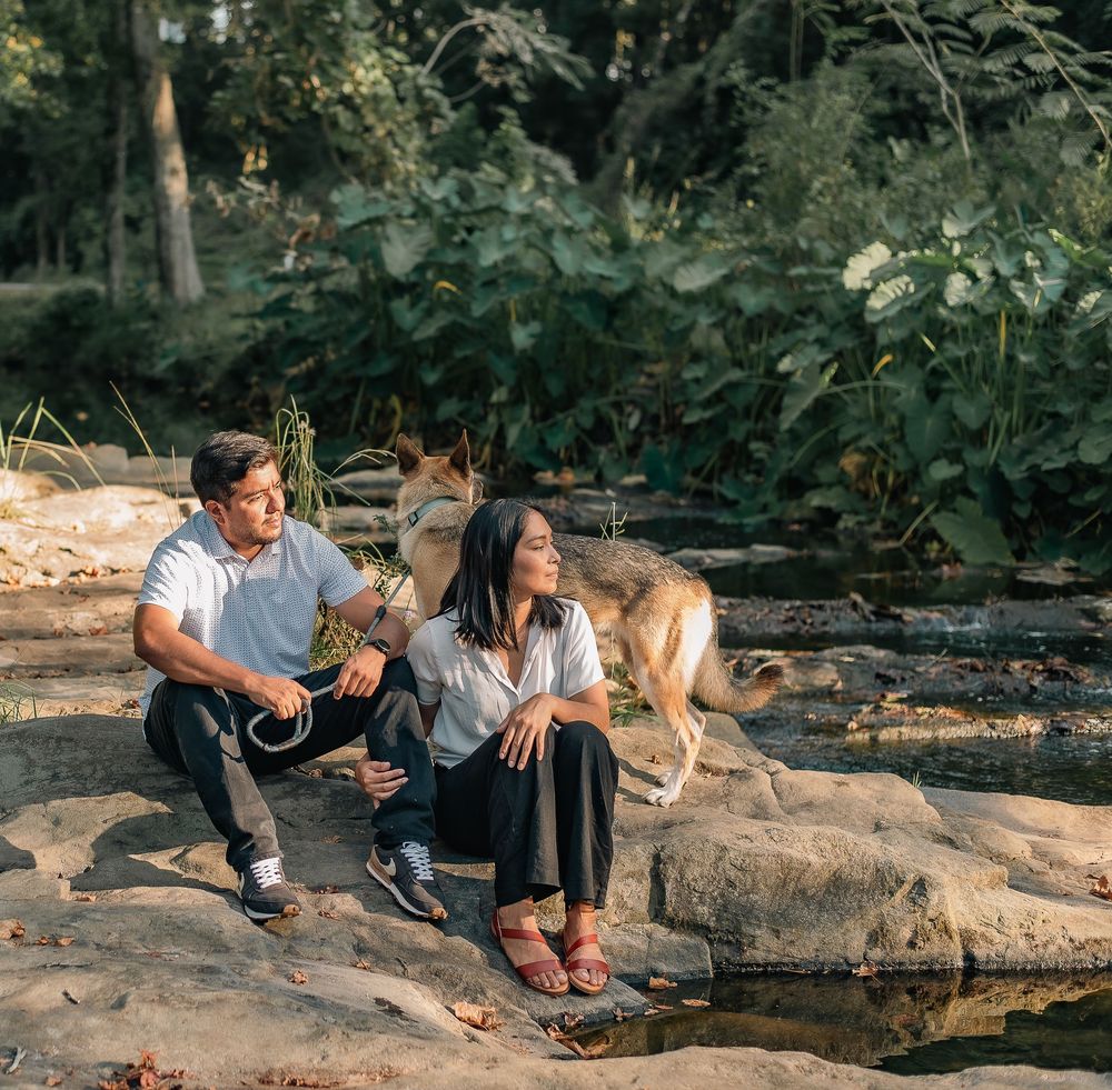 A couple and their dog enjoy a peaceful moment sitting on rocks by a stream in a wooded setting.