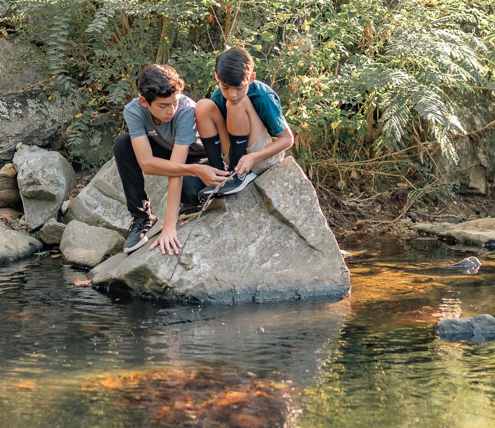 Two people sit together on a large rock overlooking a serene pond in nature.