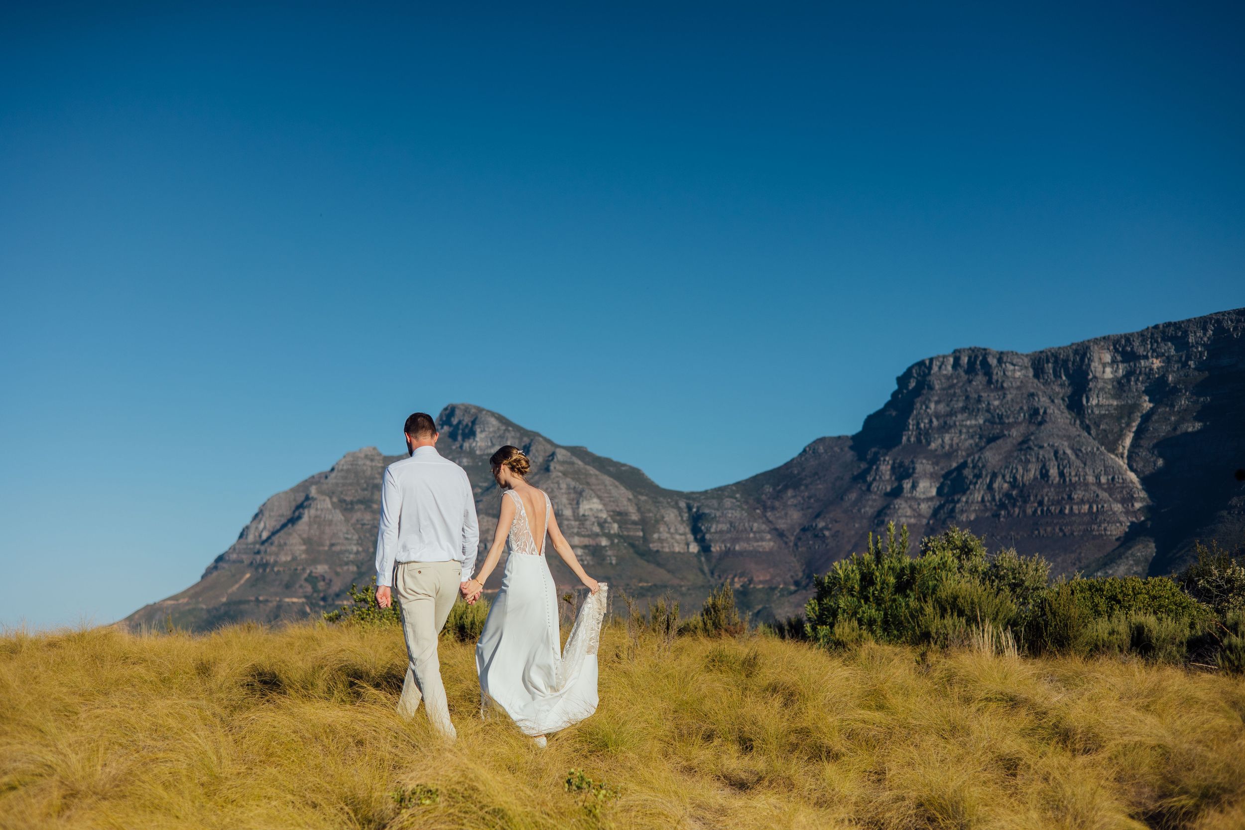 A couple stands together in a grassy field with majestic Table Mountain and clear blue skies in the background.
