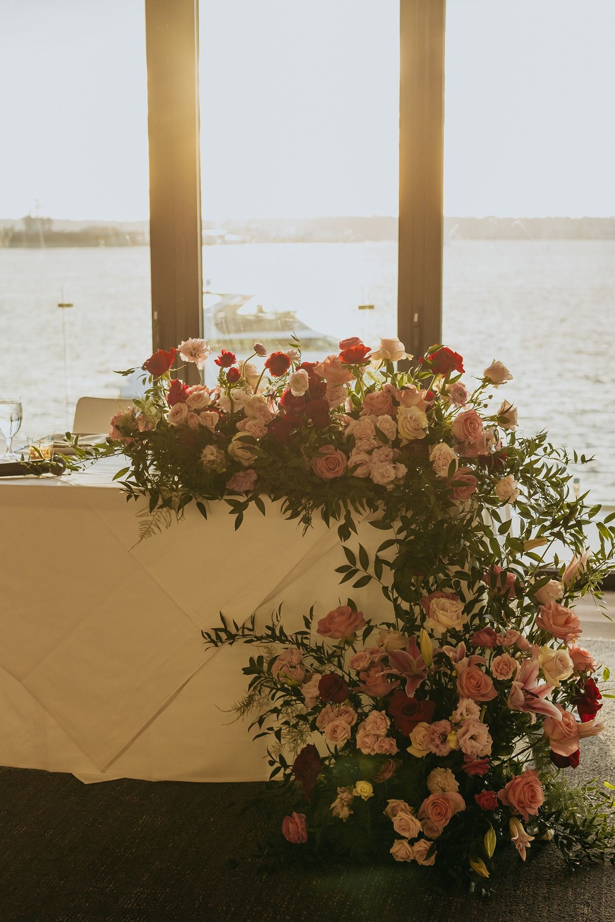 Pink floral arrangements and greenery decorate a seaside venue at sunset with ocean views through pillared windows.