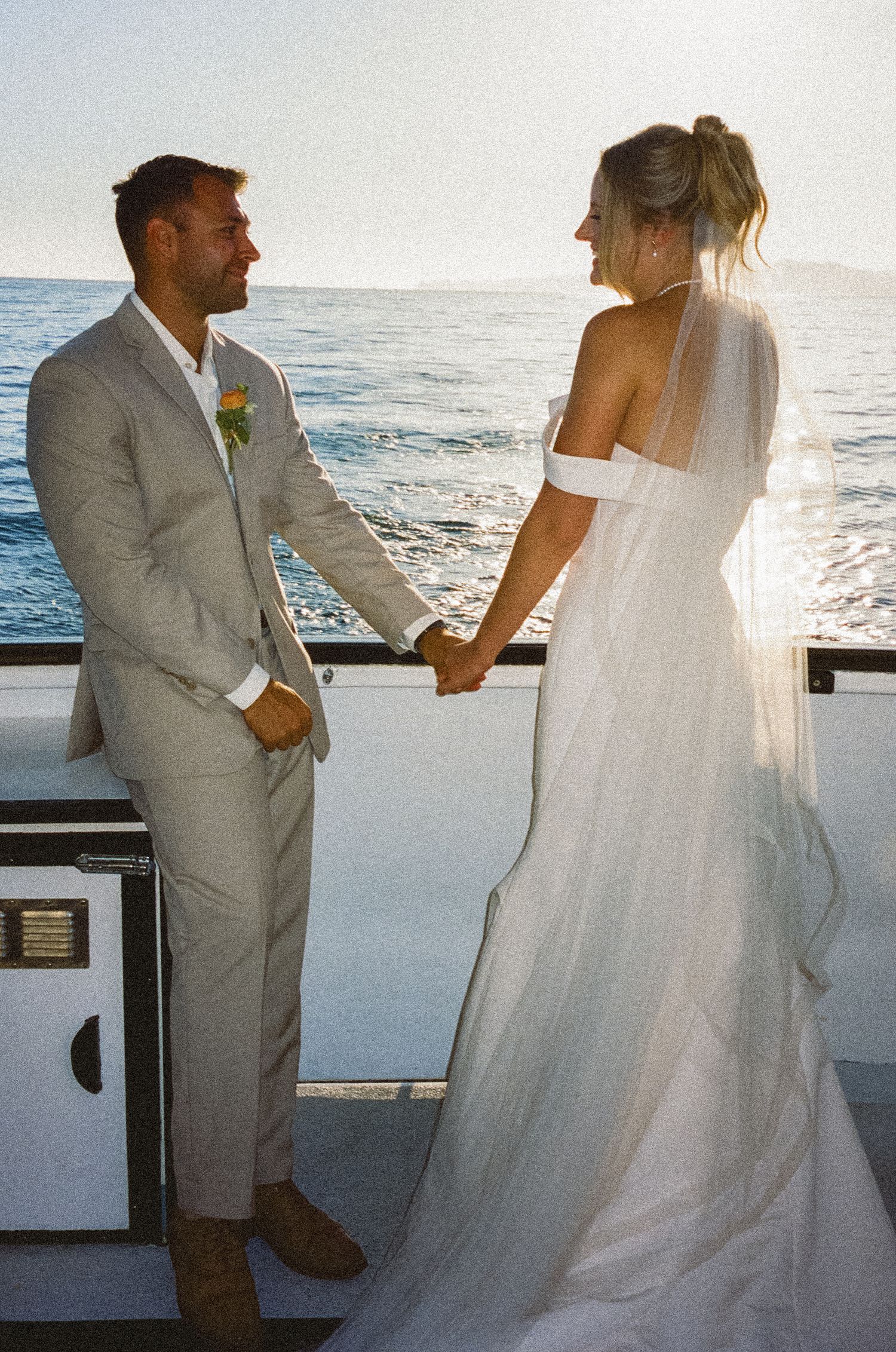 A bride and groom share a joyful moment on a boat deck at sunset, with ocean waves and glowing light in the background.