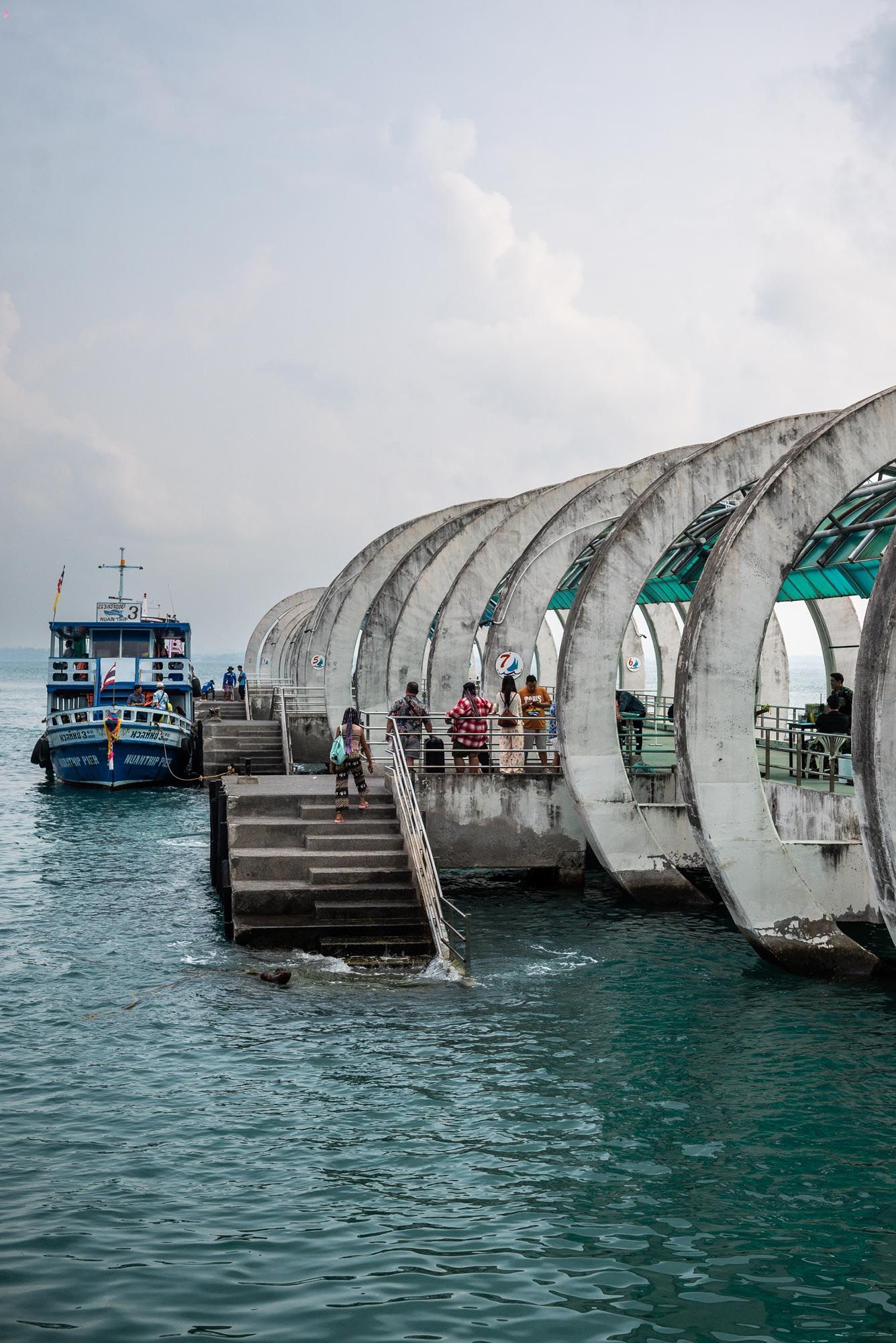 A series of curved concrete barriers form a breakwater structure extending into turquoise waters with boats visible in the distance.