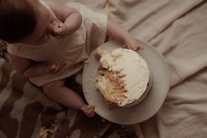 A slice of white frosted cake with crumbs sits on a gray plate against cream colored fabric.