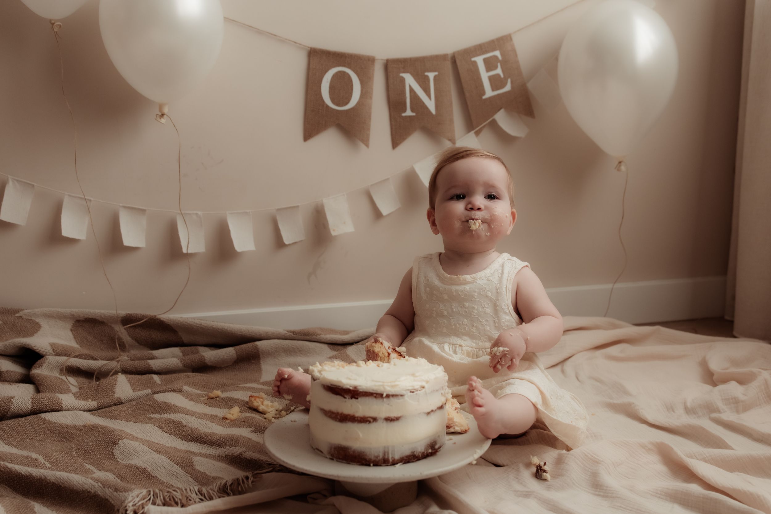 A baby sits in front of a white cake during a first birthday celebration with decorative bunting in the background.