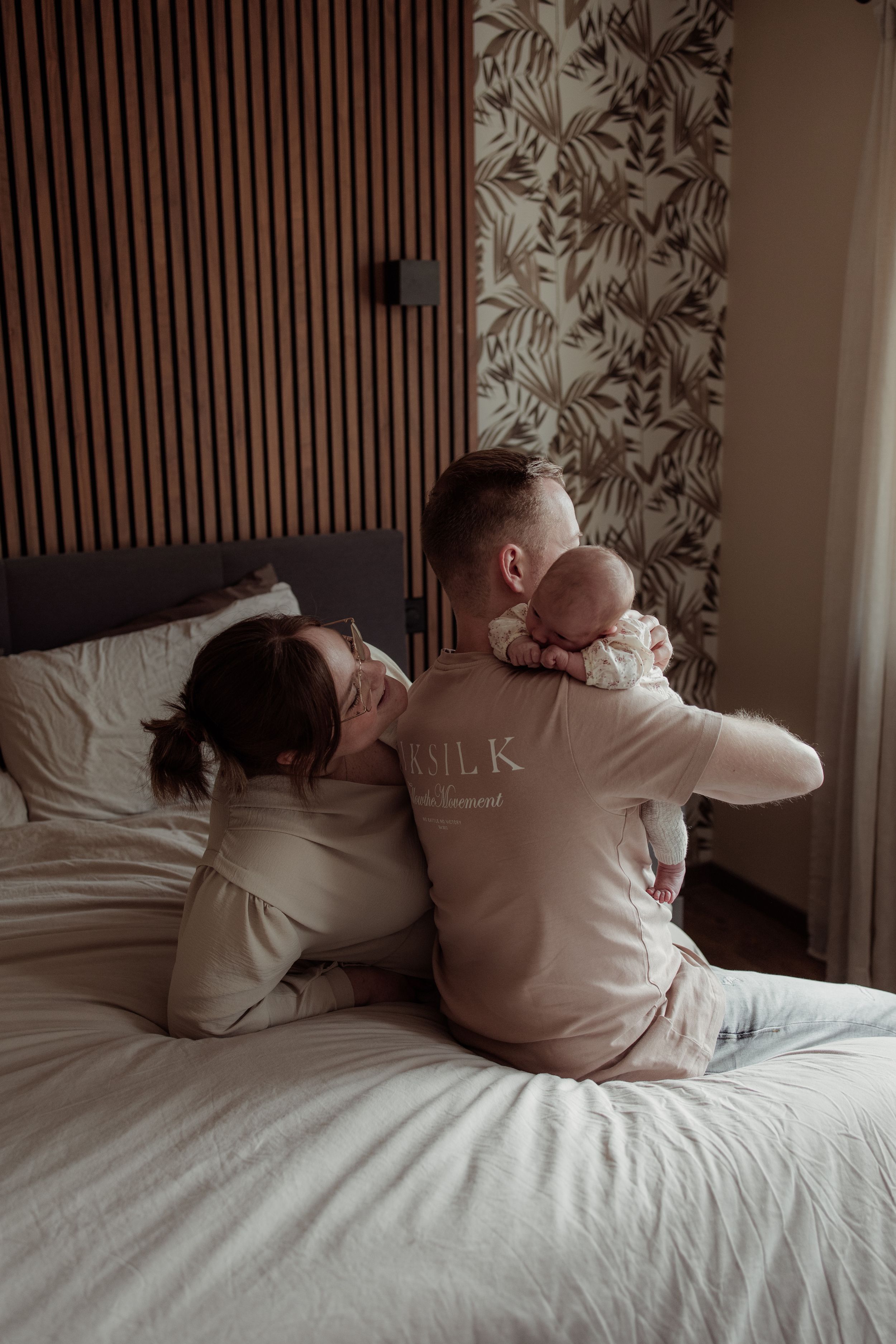 A family relaxes together on a bed with wooden slat wall decor and floral wallpaper in a cozy bedroom setting.