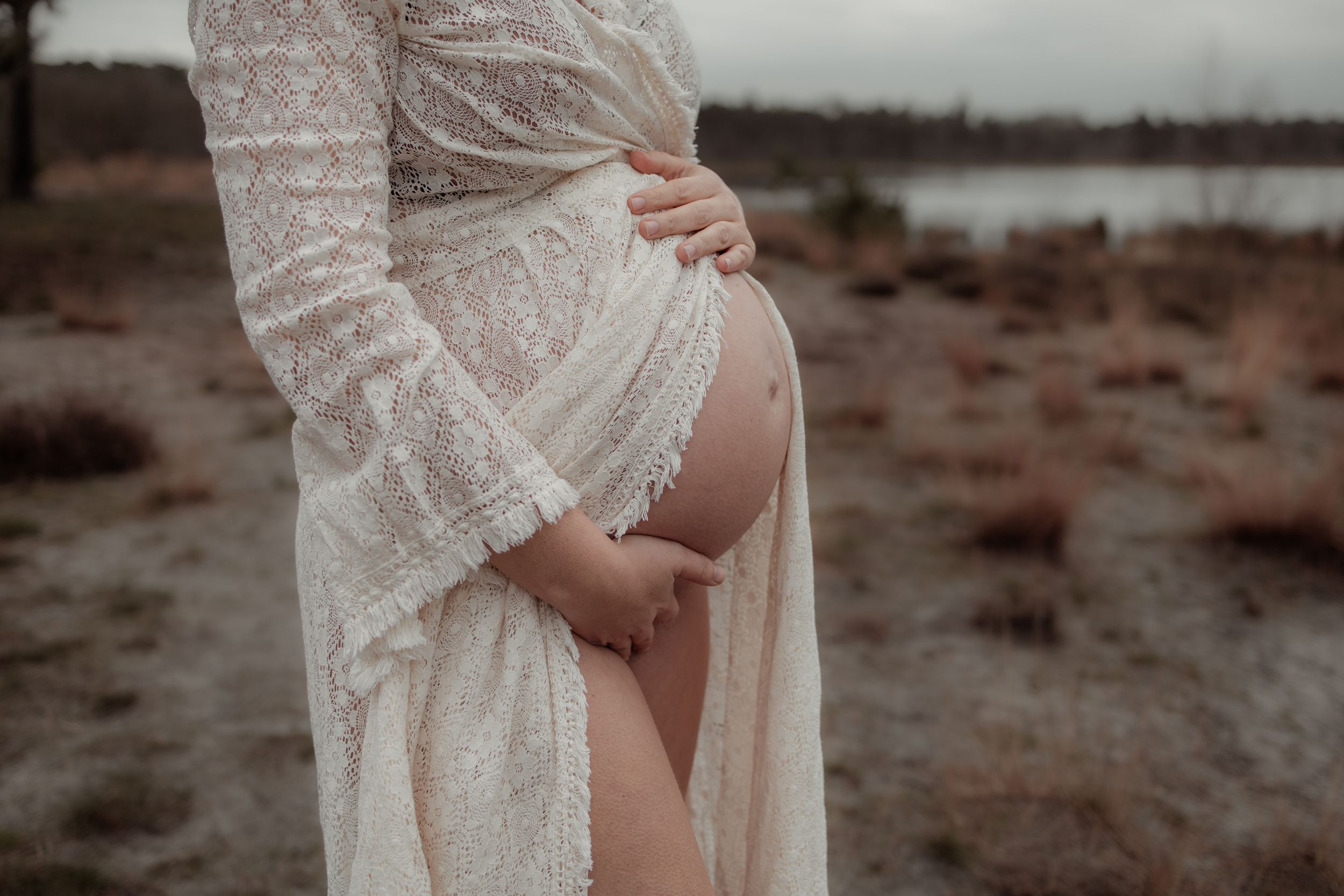 A pregnant belly draped in delicate white lace dress stands against a moody desert landscape at dusk.
