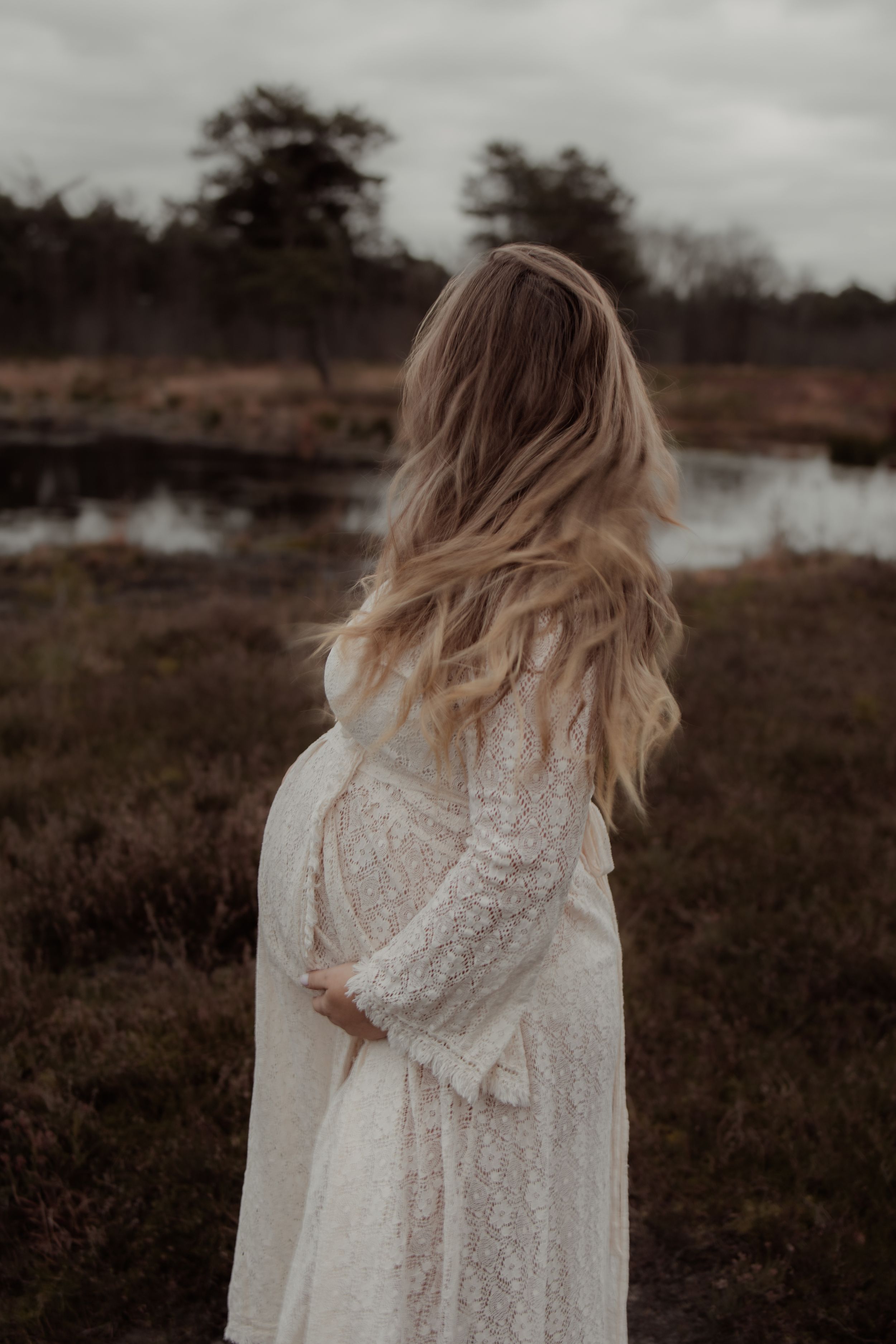 A person in a white lace dress stands in a moody field during golden hour with windswept blonde hair.