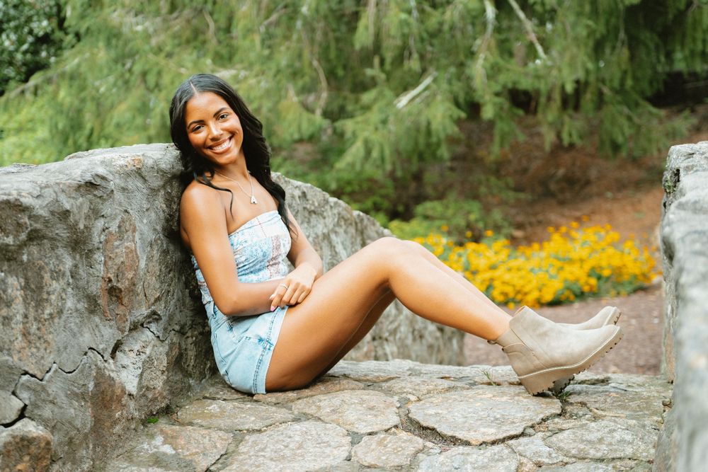 Person in denim shorts poses casually on large rocks surrounded by lush greenery and yellow flowers in outdoor setting.
