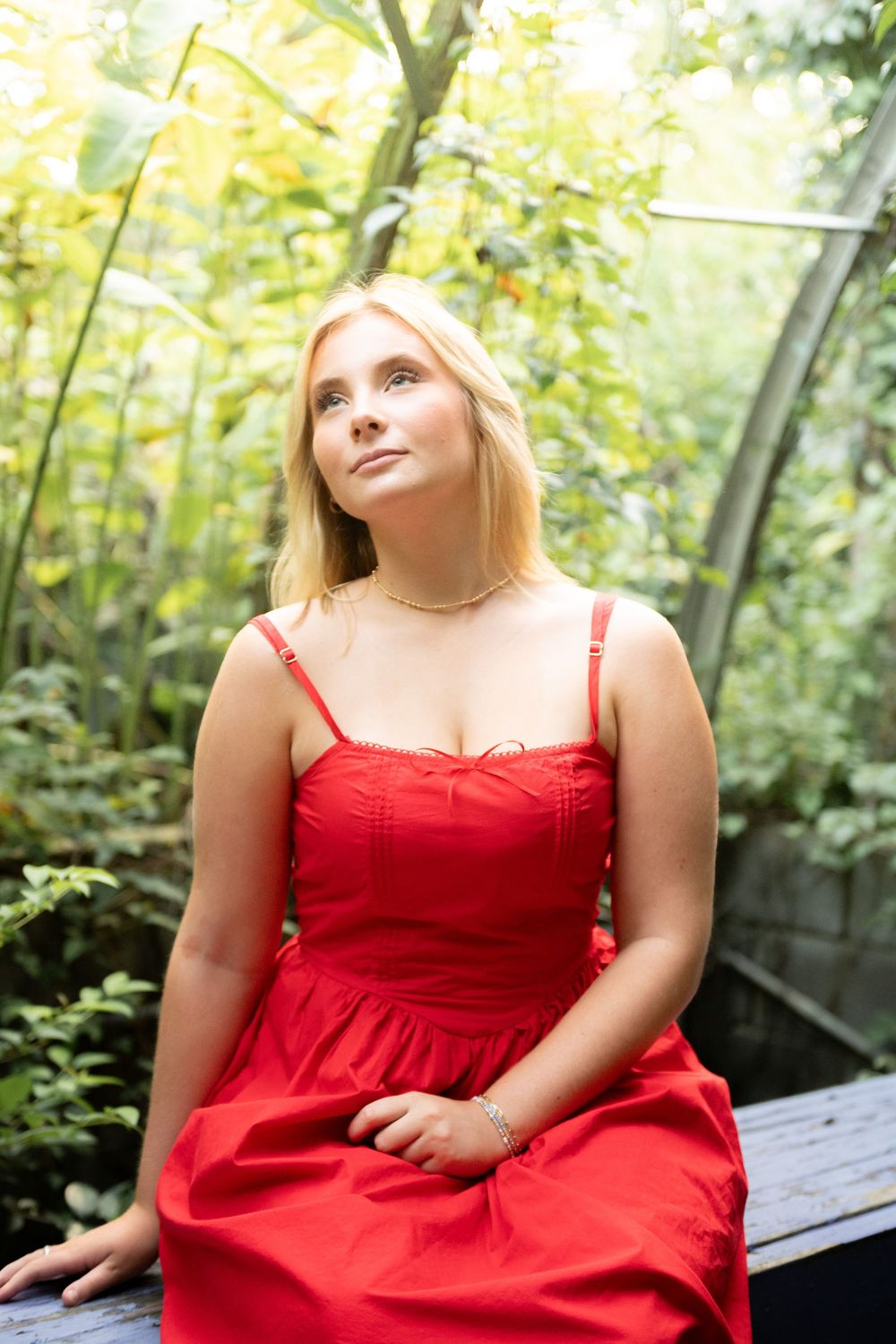 A person in a red sundress sits on a bench surrounded by lush greenery in a garden setting.