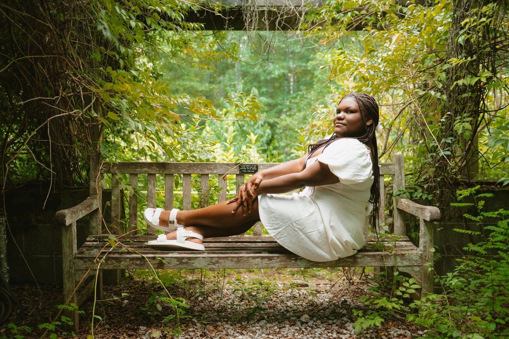 A person in a white dress lies serenely on a wooden bench surrounded by lush green forest foliage.