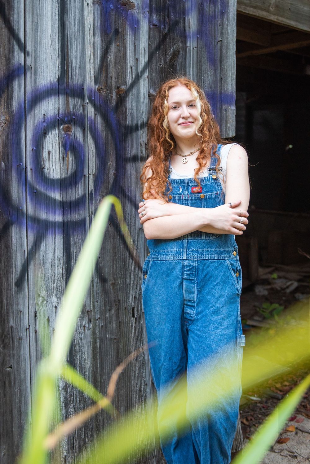 A person in denim overalls poses against a rustic wooden wall with painted swirls and blurred foliage in the foreground.