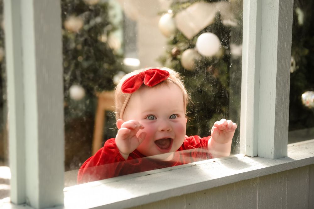 Baby in red holiday outfit crawls on white porch with Christmas decorations in background.